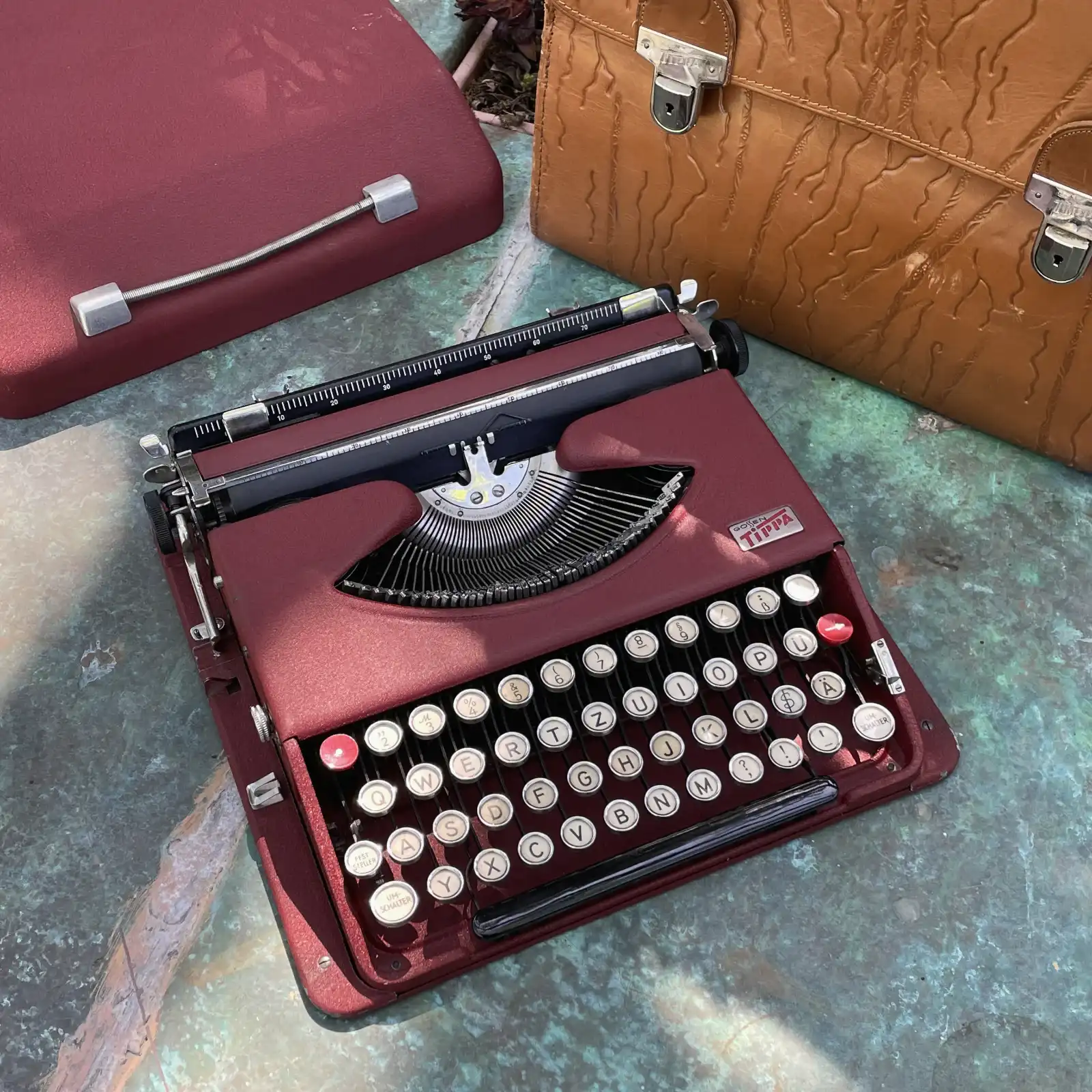 A 1948 maroon color Gossen Tippa on a table beside its lid and leather breifcase